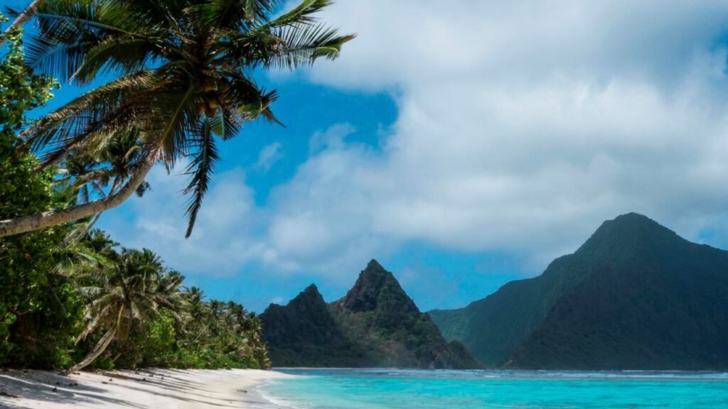 View of sandy white beach and clear turquoise waters with a backdrop of mountains at the National Park of American Samoa, credit to the National Park Service