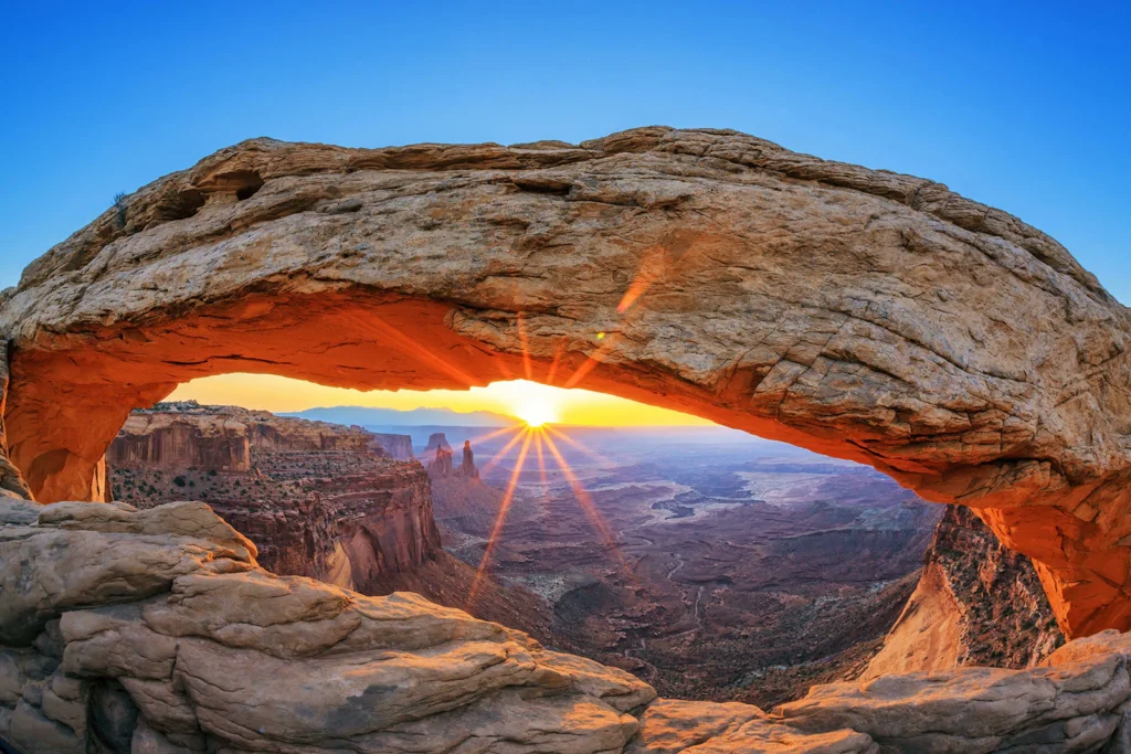 A panoramic view of a red stone arch in Canyonlands National Park on a clear blue day, overlooking the canyon and cliffs with the sun emerging underneath the arch. Credit to the National Park Foundation.